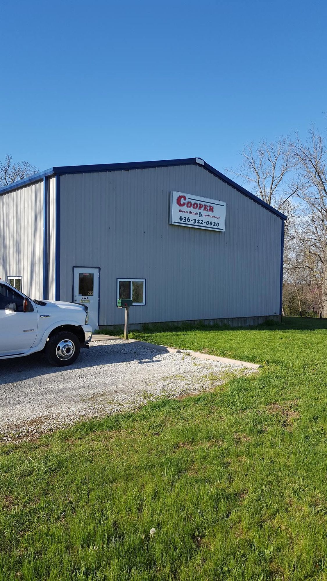 The gray metal shop building with a white pickup parked out front on the gravel