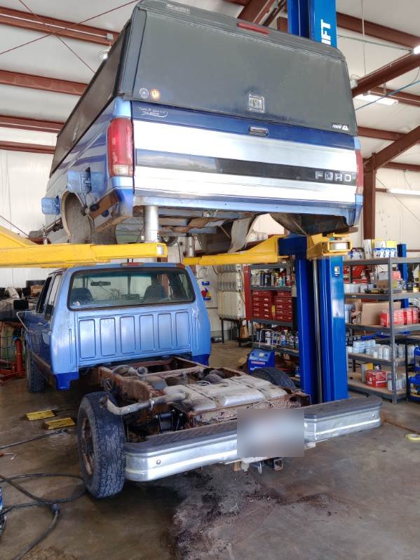 Two pickups in the shop, one raised high on a two post lift with parts shelving behind